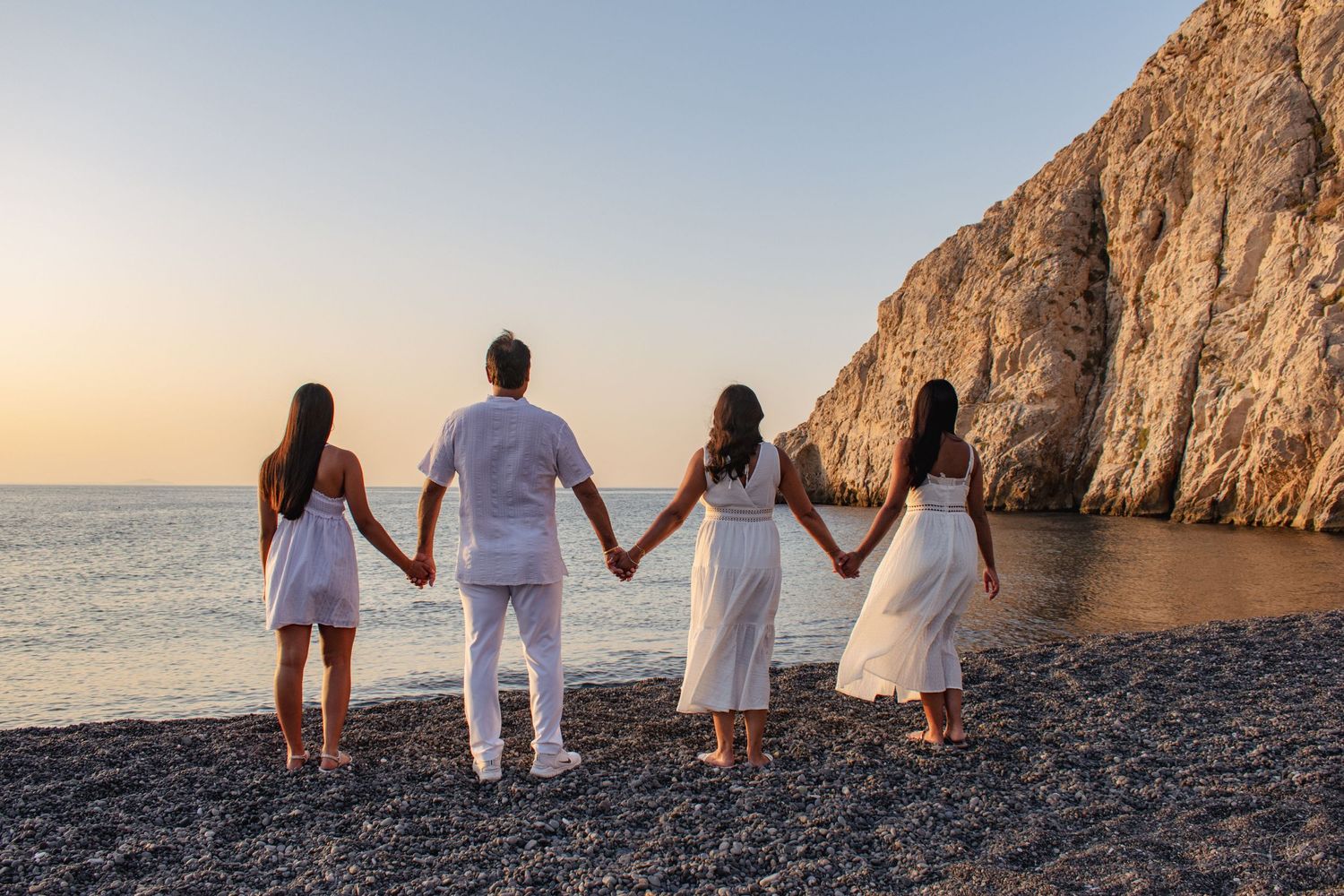Four people in white outfits hold hands while facing scenic coastal cliff view.