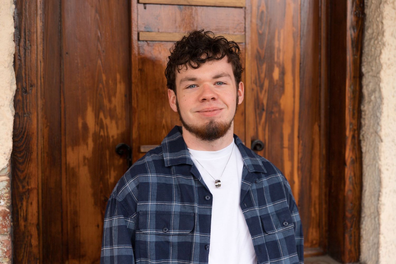 Casual portrait against wooden door wearing blue plaid flannel and white t-shirt.