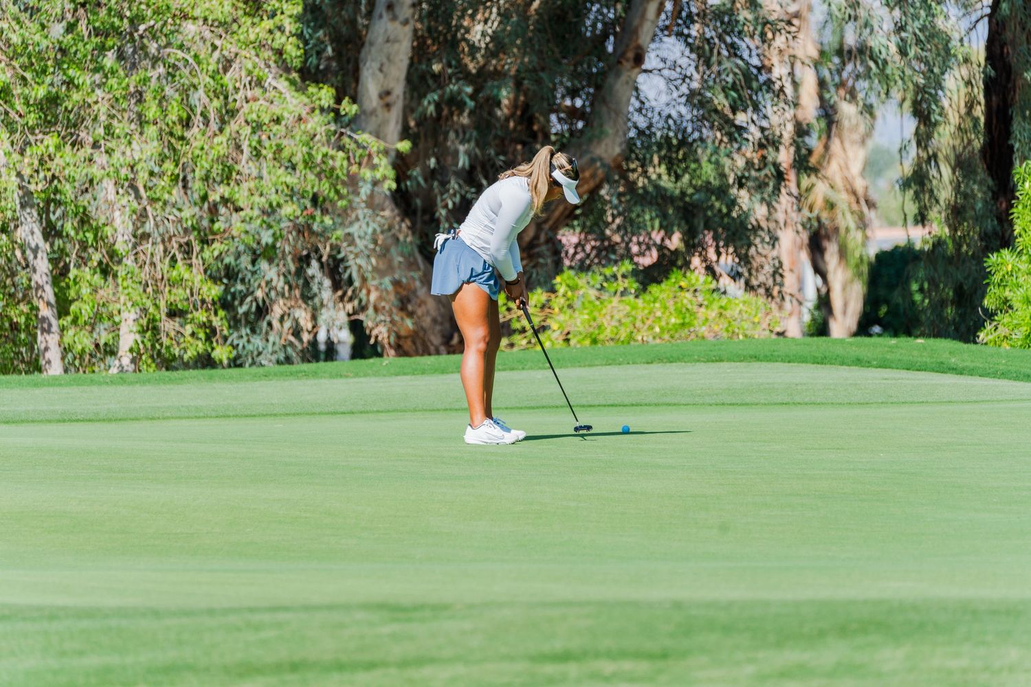Golfer in blue shorts and white top lining up a putt on a sunny golf course green.