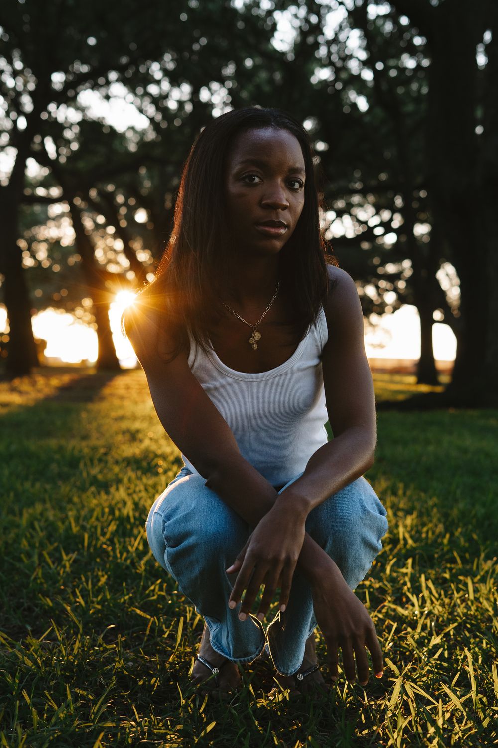 Person in white tank top sits in grass at sunset as golden sunlight filters through trees in background.