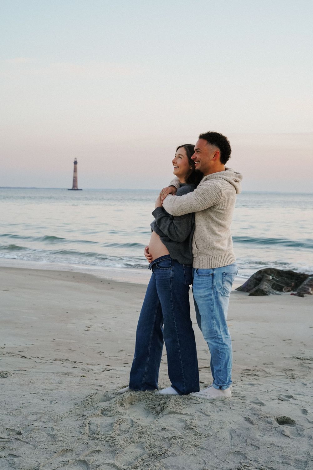 A couple embraces on a sandy beach at sunset with a lighthouse visible in the distance across multiple frames.