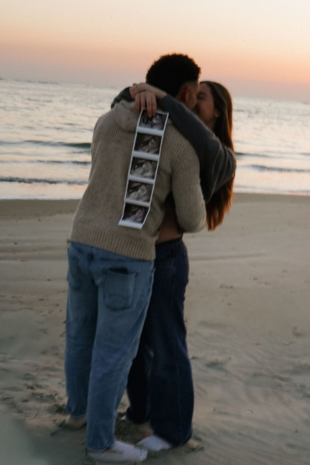 A couple embraces on a sandy beach at sunset while holding ultrasound photos.