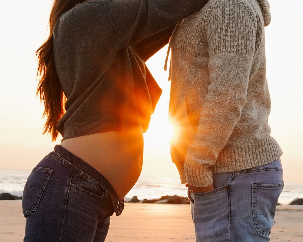Couple holds hands at sunset on a beach with golden sunlight streaming through their silhouettes.