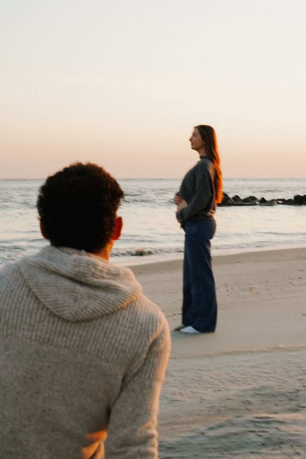 A person in a sweater sits on the beach watching another figure standing by the ocean at sunset.