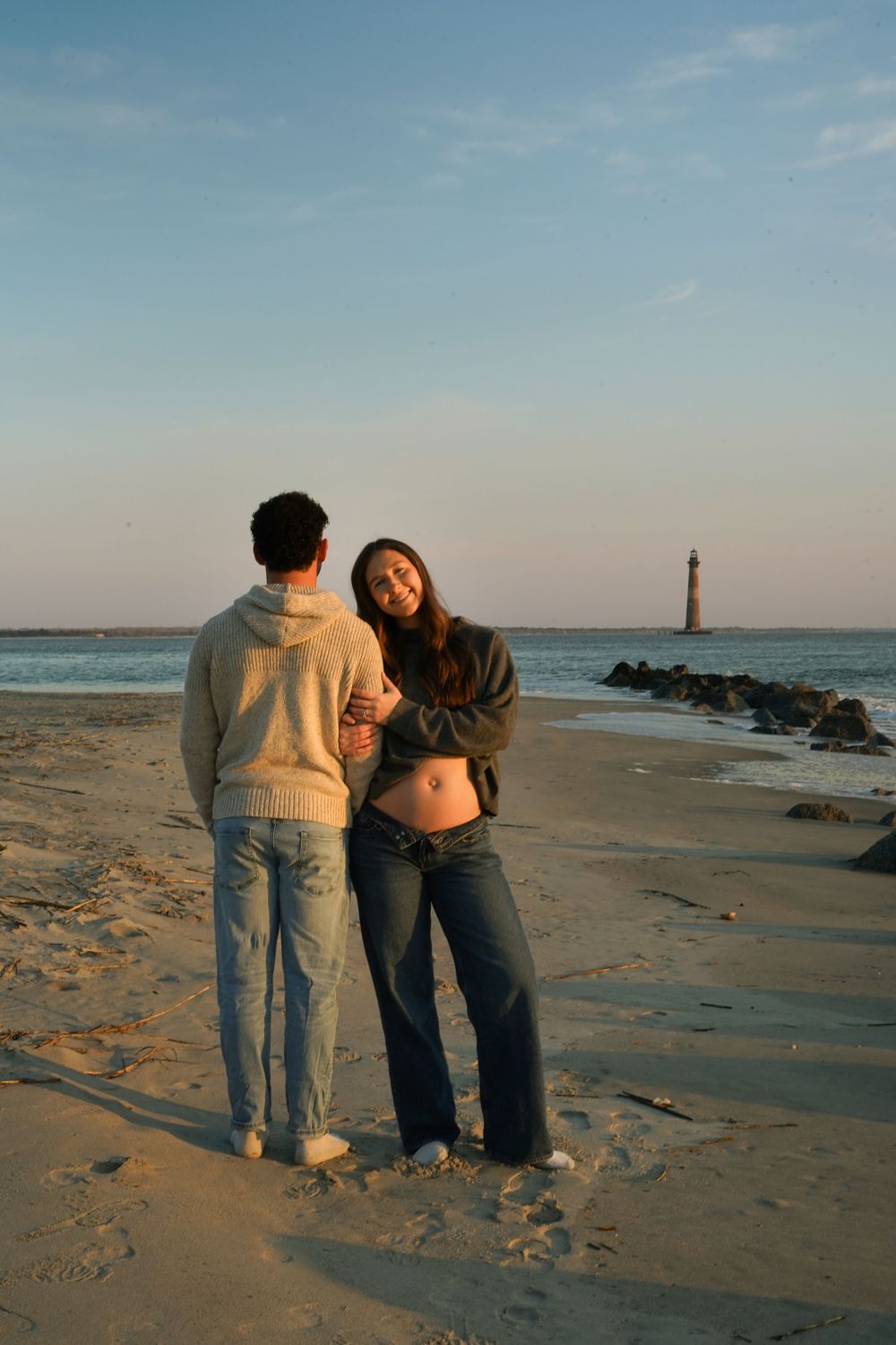 Two people bend down to examine something on a sandy beach at sunset with a lighthouse visible in the distance.