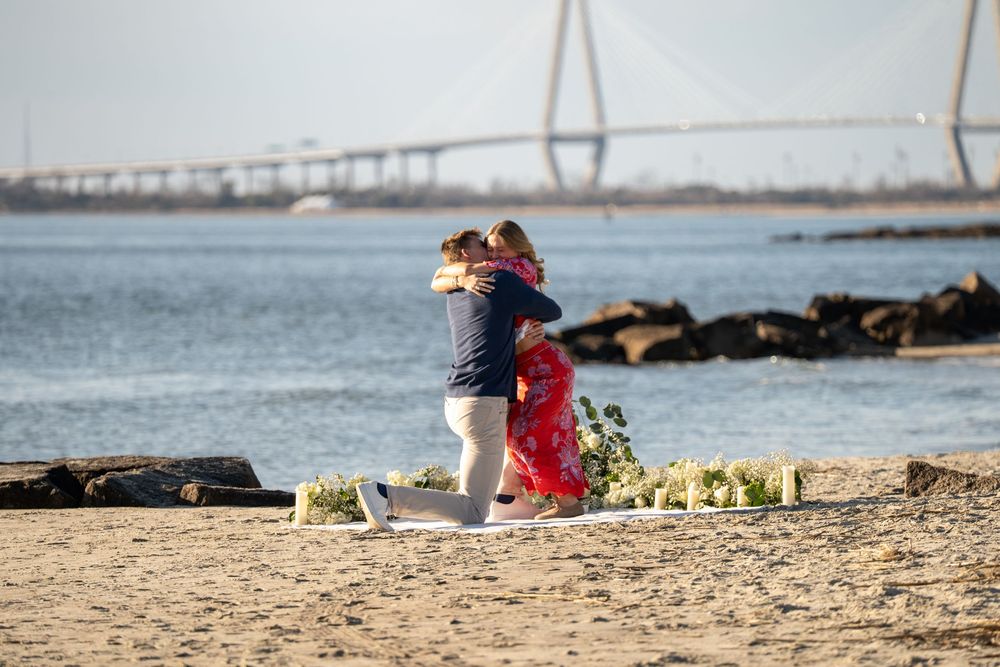 Two people embrace while standing on a beach with the Ravenel Bridge visible in the background in Charleston, South Carolina.