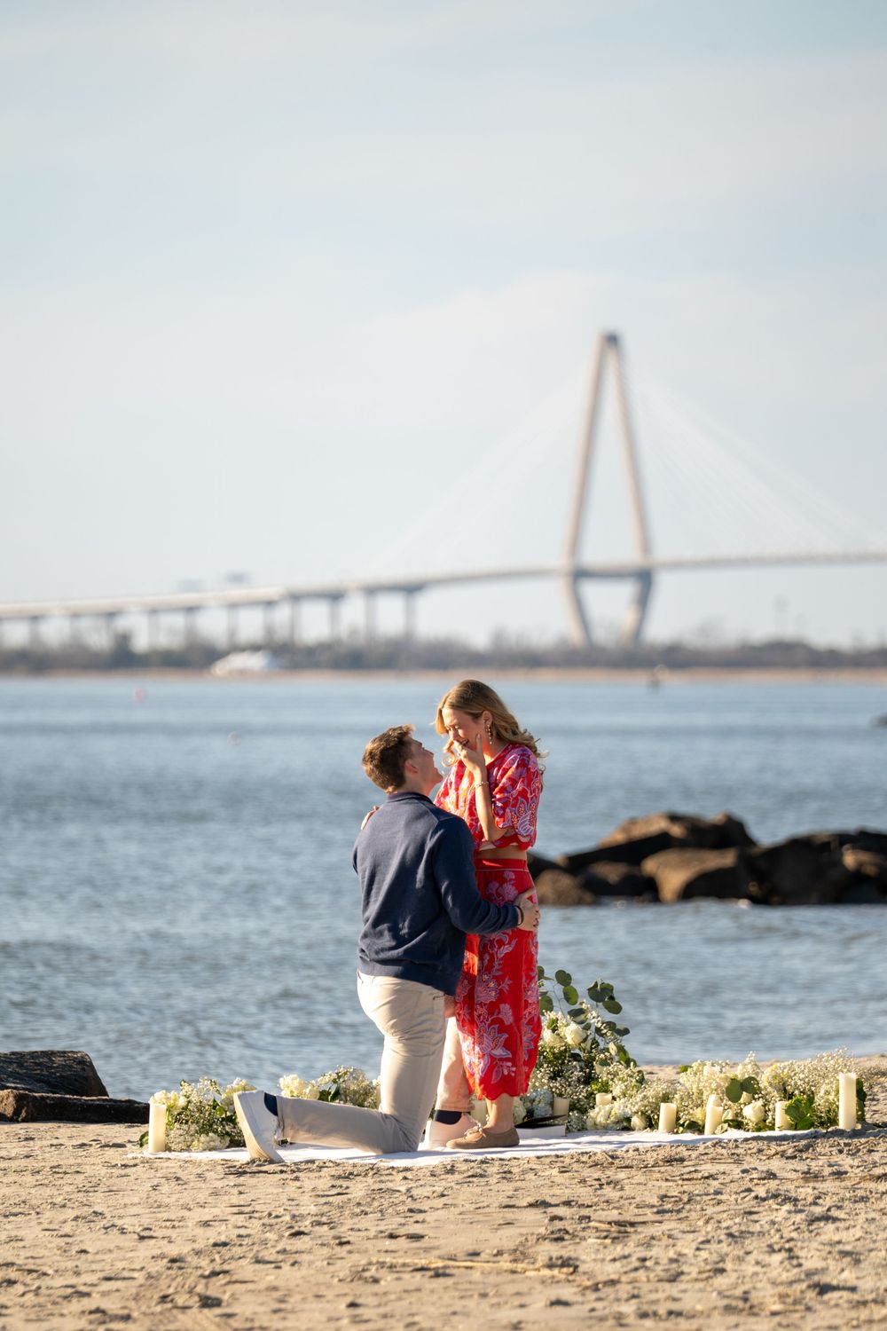 A romantic proposal scene on a beach with the Cooper River Bridge in Charleston, South Carolina in the background.