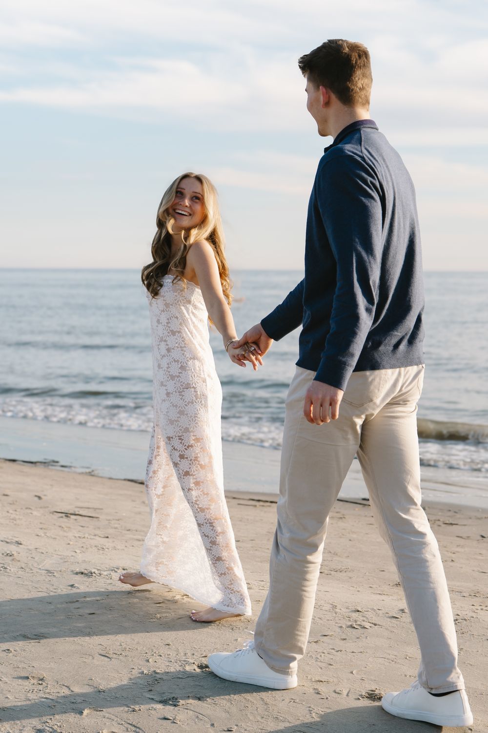 A couple in white and navy outfits dances playfully on a sunny beach with ocean waves in the background.