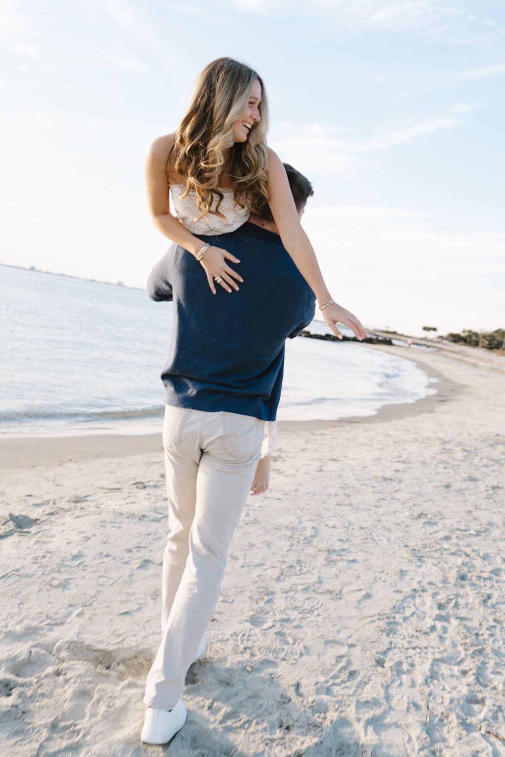 A couple in casual beach attire walks together on white sand with the ocean and blue sky in view.