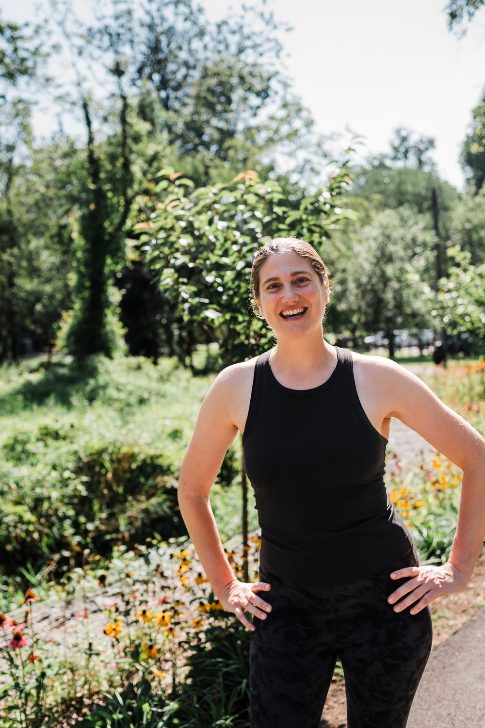 Someone in a black sleeveless top poses playfully in a sunny garden setting with flowers and greenery.
