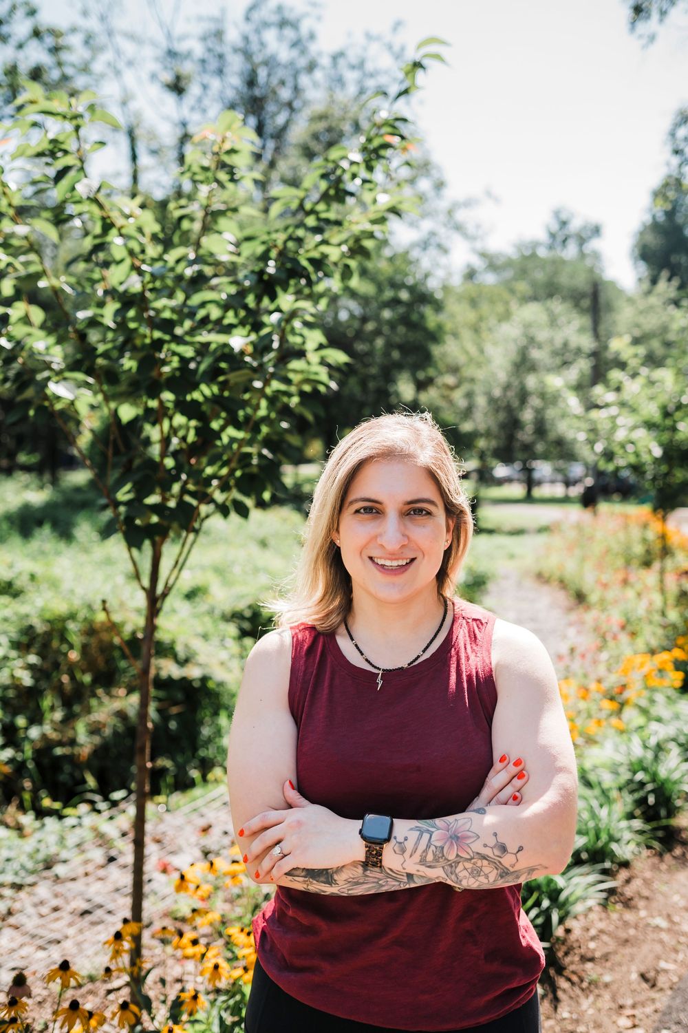 A person in a burgundy sleeveless top poses in a sunny garden filled with flowers and small trees.