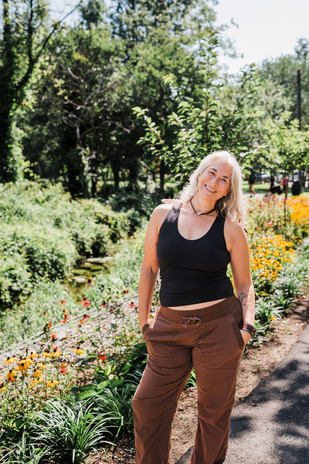 A paved walking path through a lush garden with trees and flowering plants in summer.