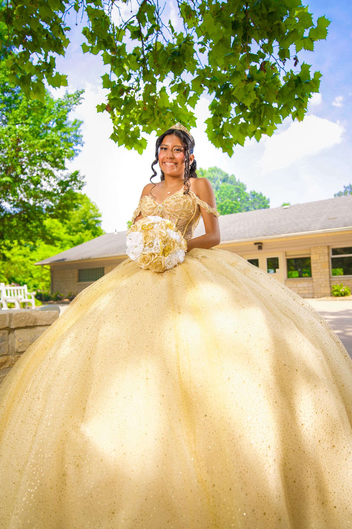 QUINCEAÑERA gold ballgown with sparkles photographed outdoors under green trees and blue sky.