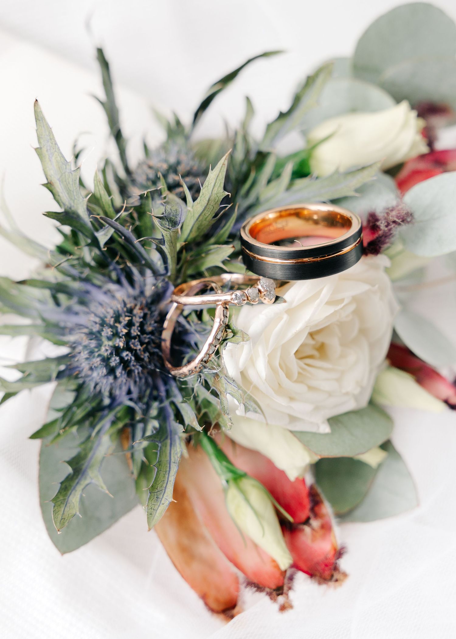 Wedding rings displayed on white roses and blue thistle with eucalyptus leaves.