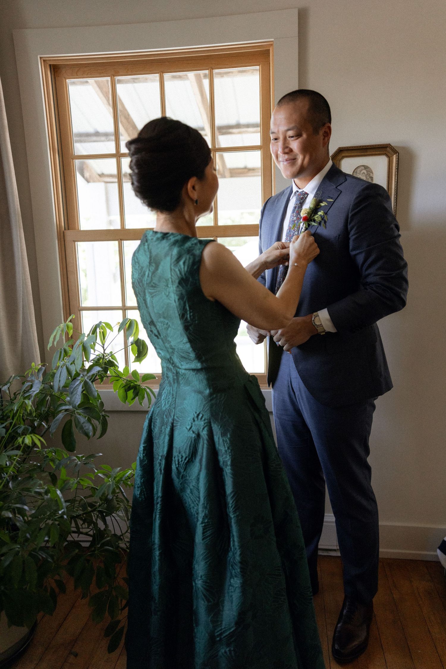 A formal dress fitting scene in a sunlit room with houseplants near windows.