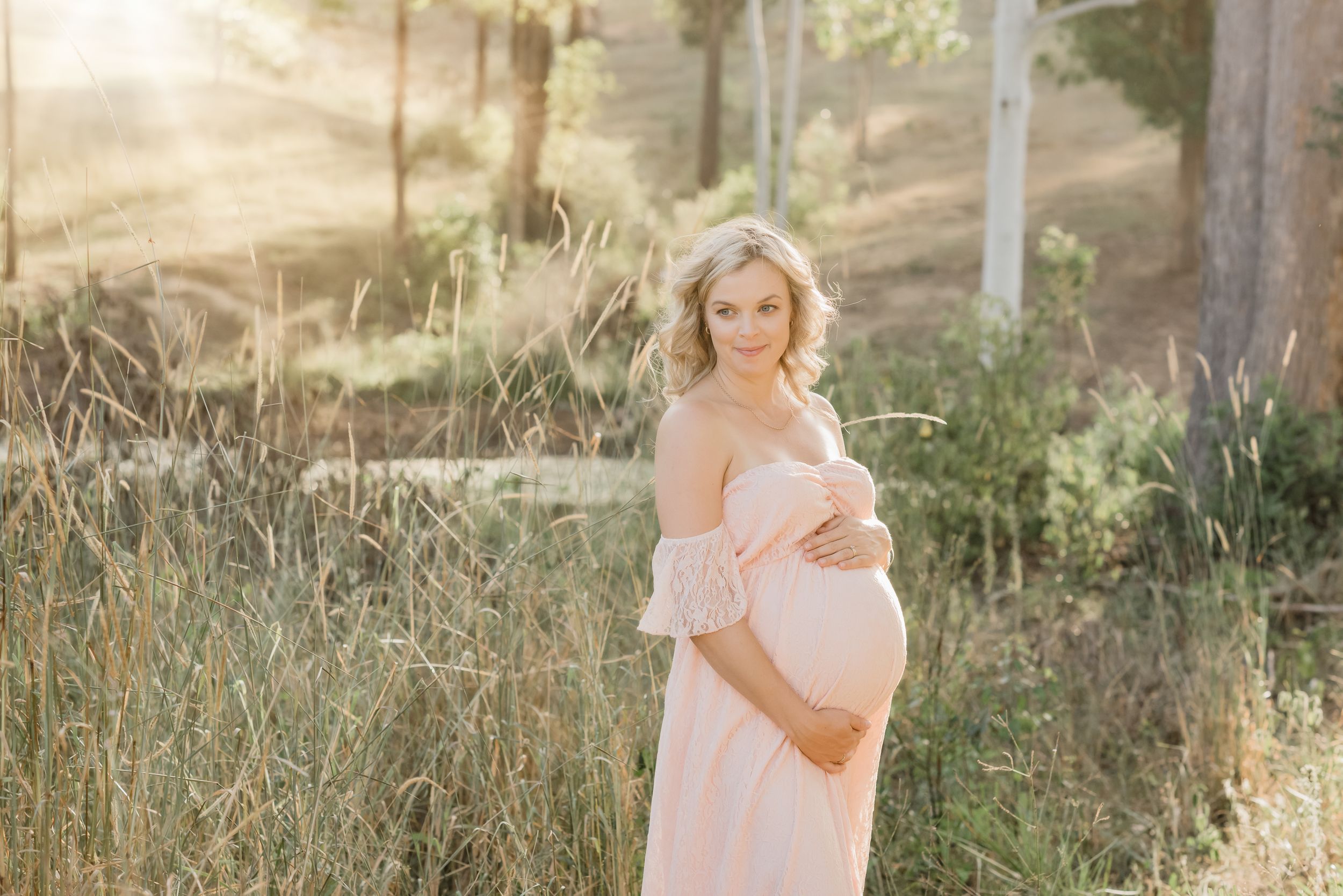 Maternity photo of woman in flowing pink dress standing in golden sunlit field.