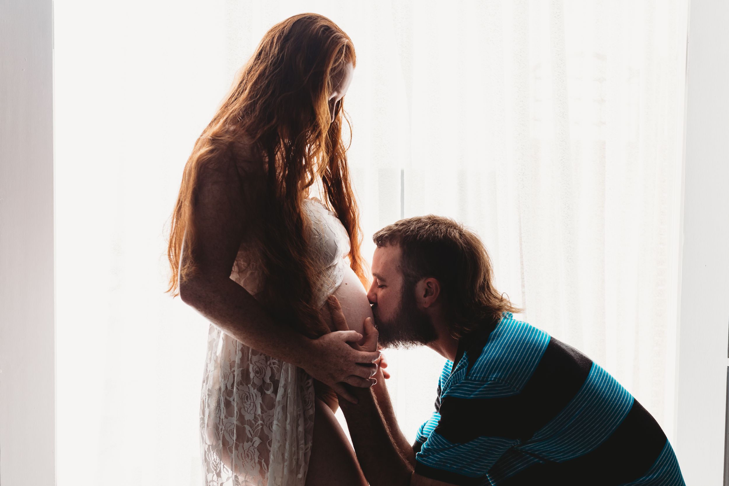 An intimate maternity moment captured in soft window light with a white flowing dress.