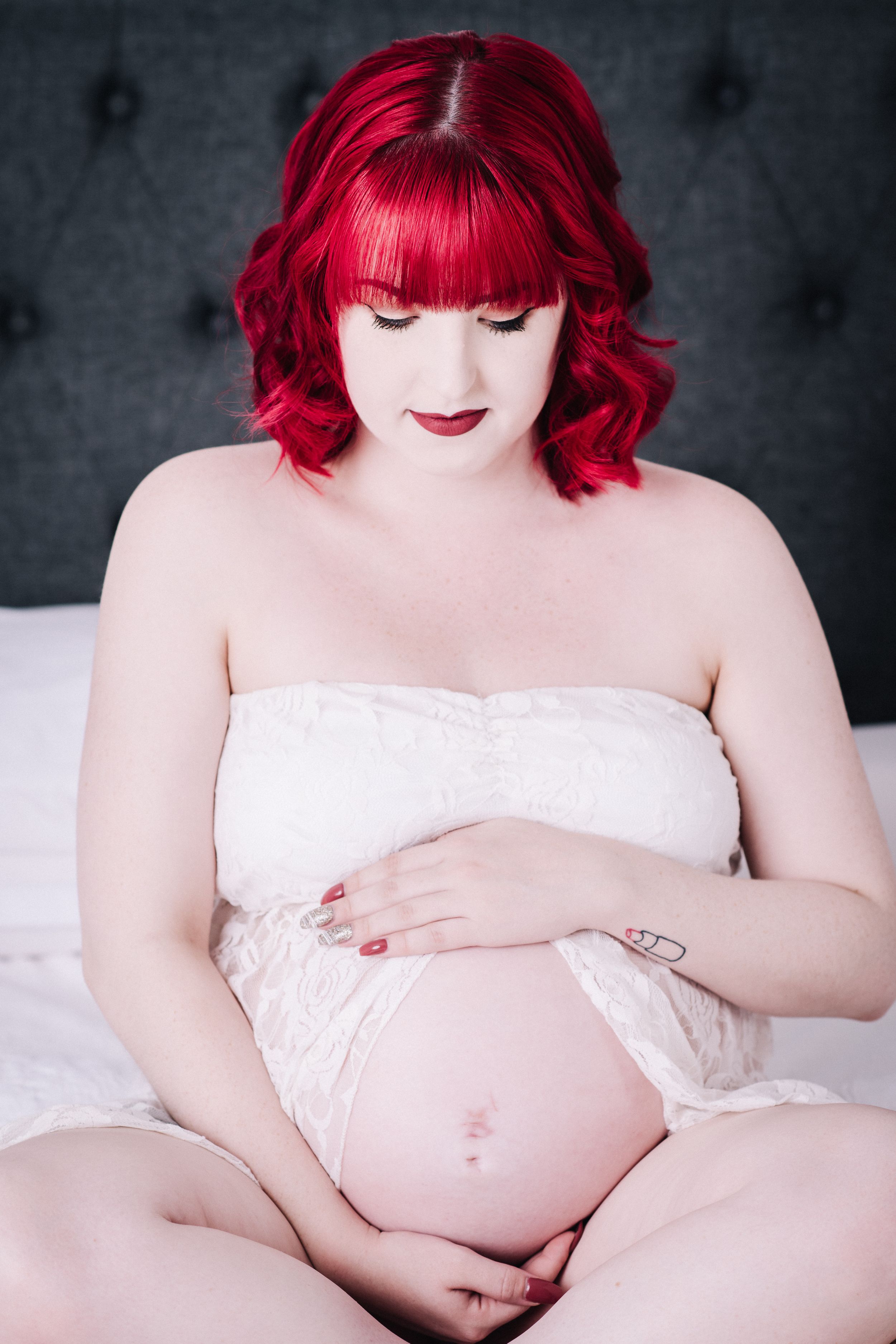 Maternity portrait featuring woman with red hair in white lace against dark background.