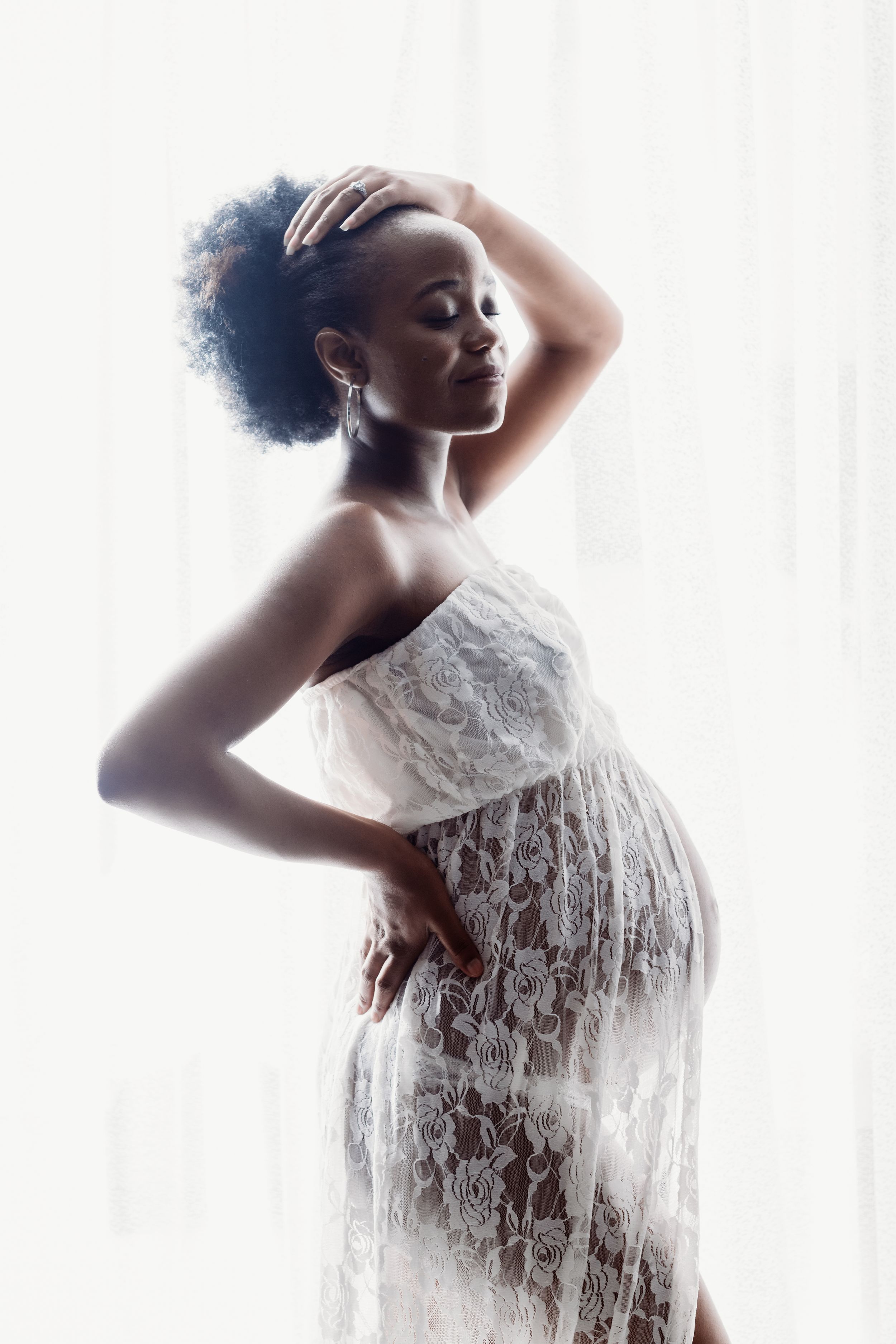 An artistic maternity portrait featuring a woman in white lace dress with backlit window glow.