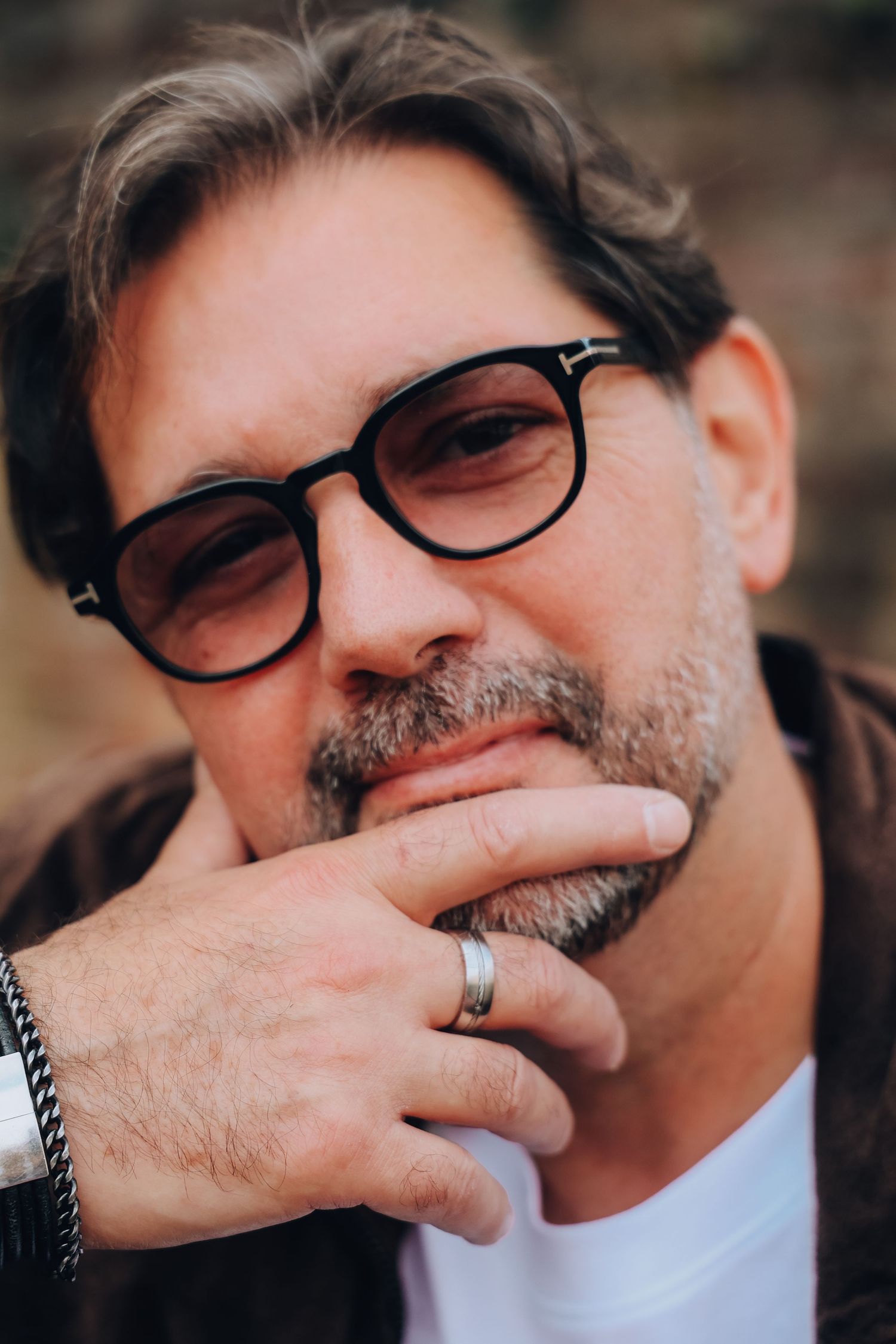 A mature man with glasses and a silver ring rests his chin on his hand in a close-up portrait.