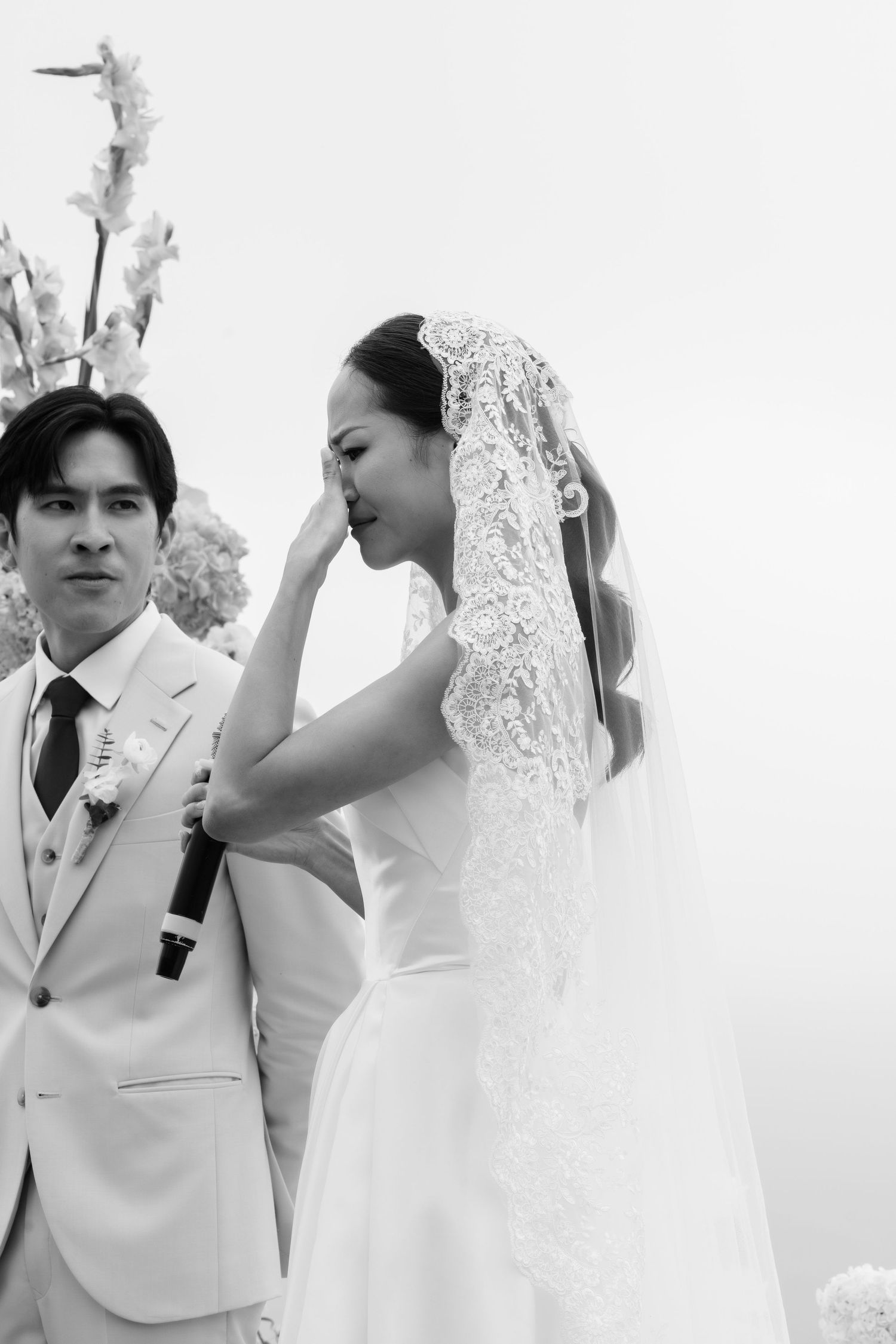Emotional bride wiping tears during outdoor wedding ceremony beside groom in beige suit, black and white photo.