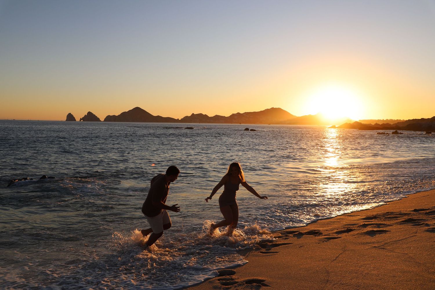 Romantic beach photo of couple in white clothing during golden hour with waves crashing behind.