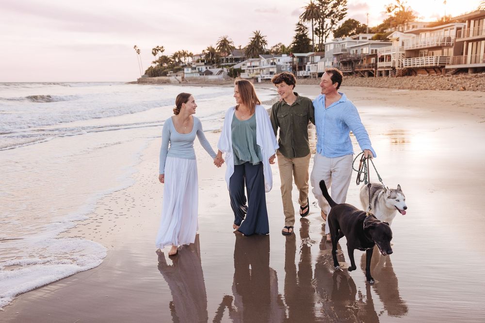 Friends stroll along the wet sand of a beach at golden hour with their dogs beside them.