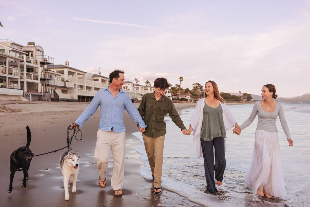 People walk their dogs along a scenic beach with coastal homes visible in the background.