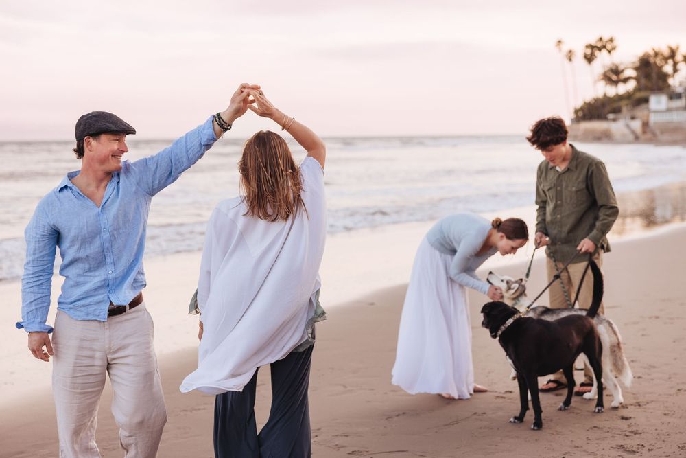 People enjoying a sunset walk with their dog along a sandy beach while taking photos on a peaceful evening.