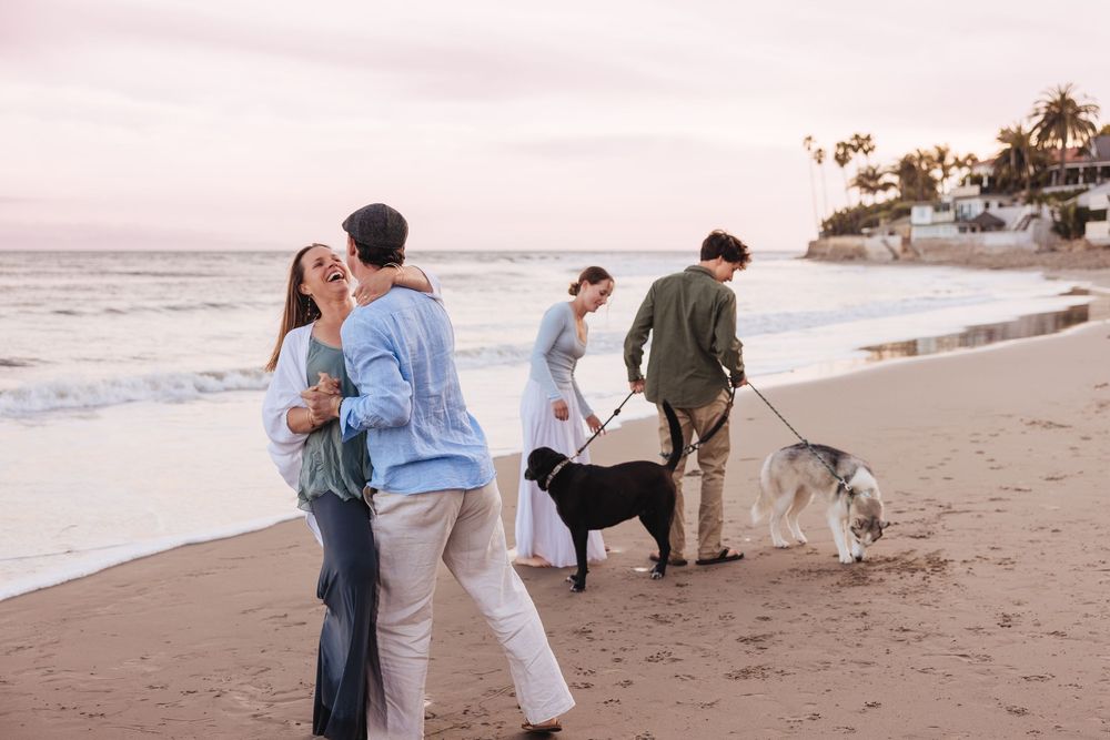 Loving couple with their black dog taking engagement photos on a sandy beach at golden hour.