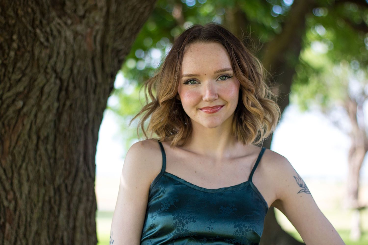 A person in a teal blue dress poses outdoors near a tree while gently smiling at the camera.