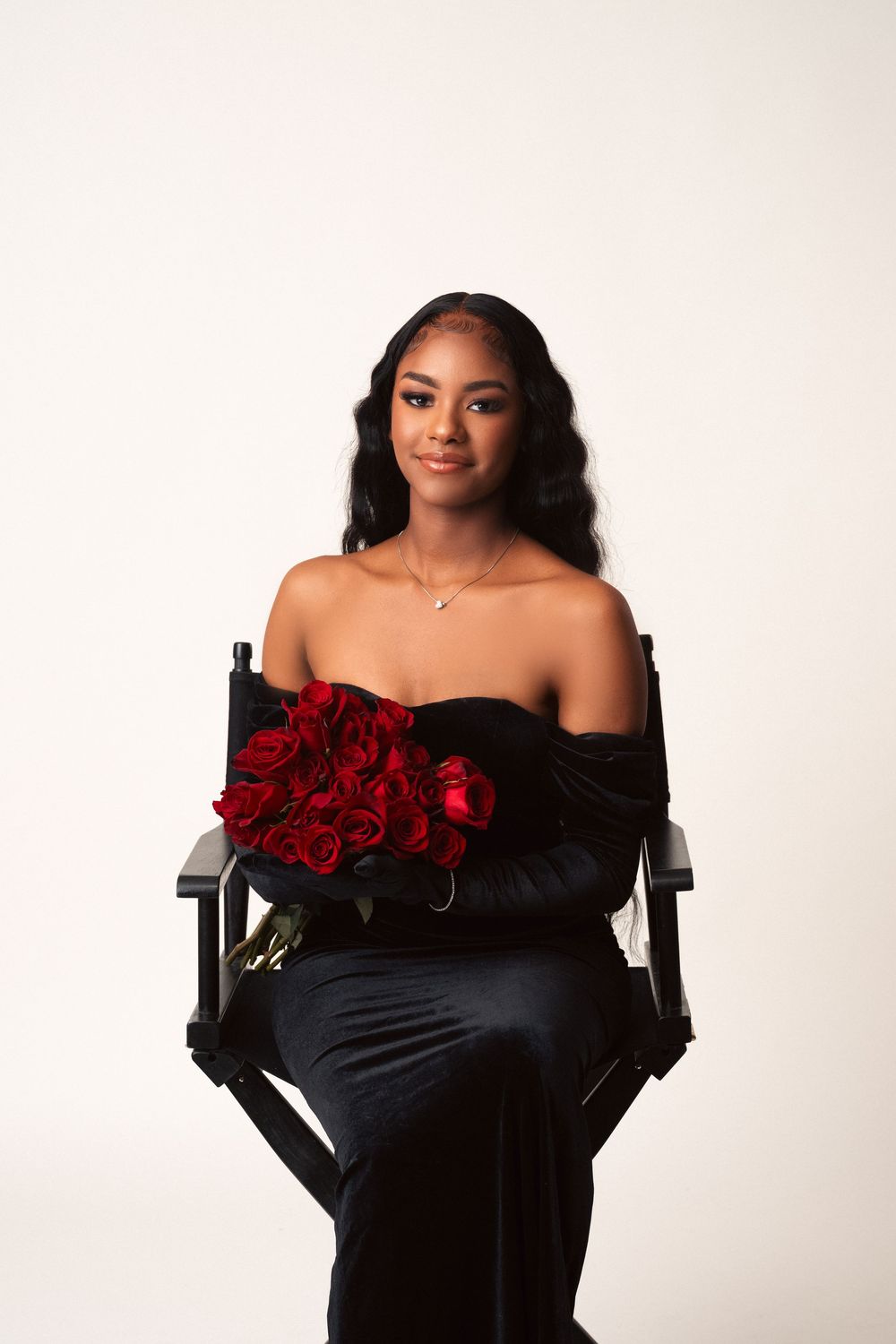 Elegant portrait of someone in a black off-shoulder dress holding red roses while seated in a director's chair against white backdrop.