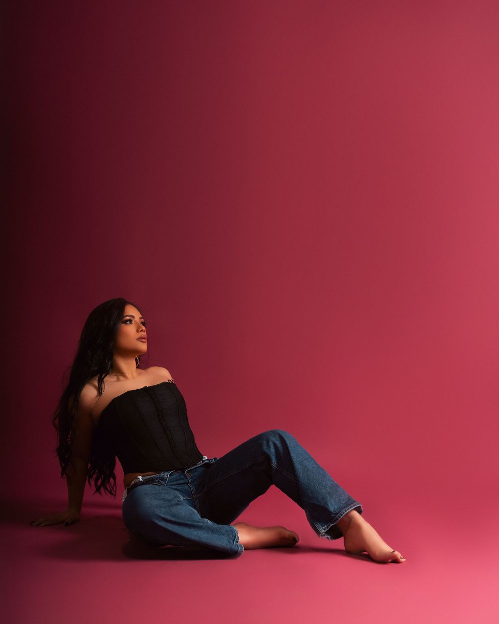 A person in a black top and jeans reclines on the floor against a vibrant pink studio backdrop.