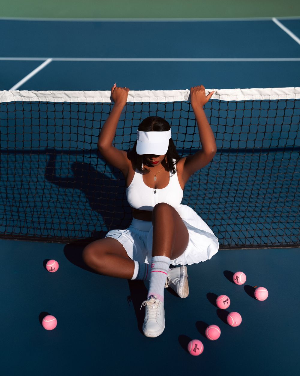 Tennis player in white outfit lying on court with pink tennis balls scattered around on blue hard court surface.
