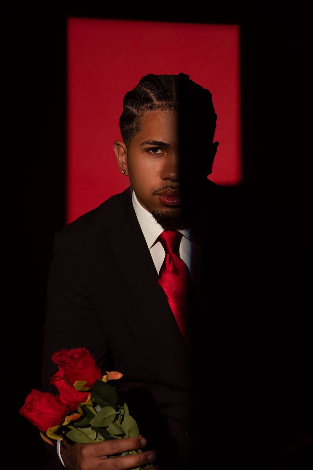 Dramatic portrait against red backdrop shows figure in dark suit with red tie holding red roses in moody lighting.