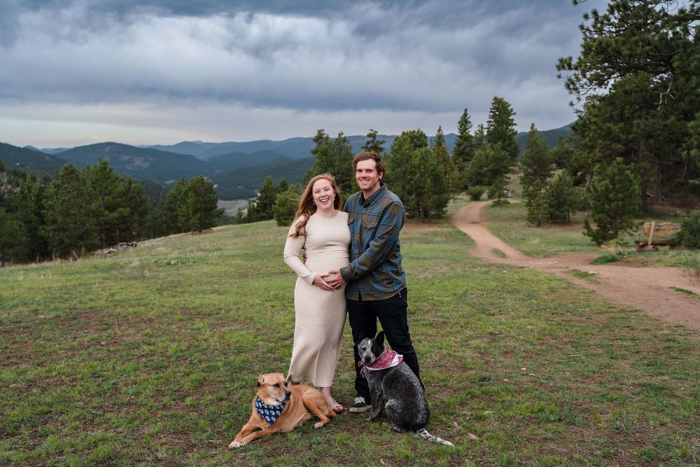 A couple sits together on a hillside overlooking scenic mountain vistas with dramatic cloudy skies in the background.