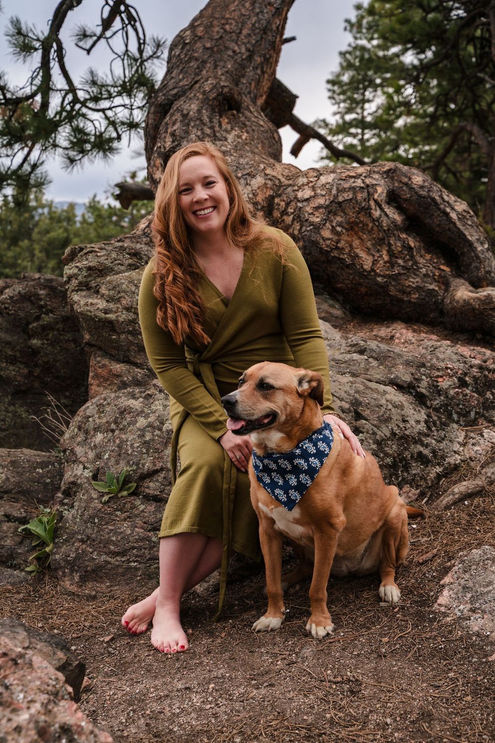 Person in olive dress sits with a brown and white bulldog near a large gnarled tree trunk in a natural outdoor setting.