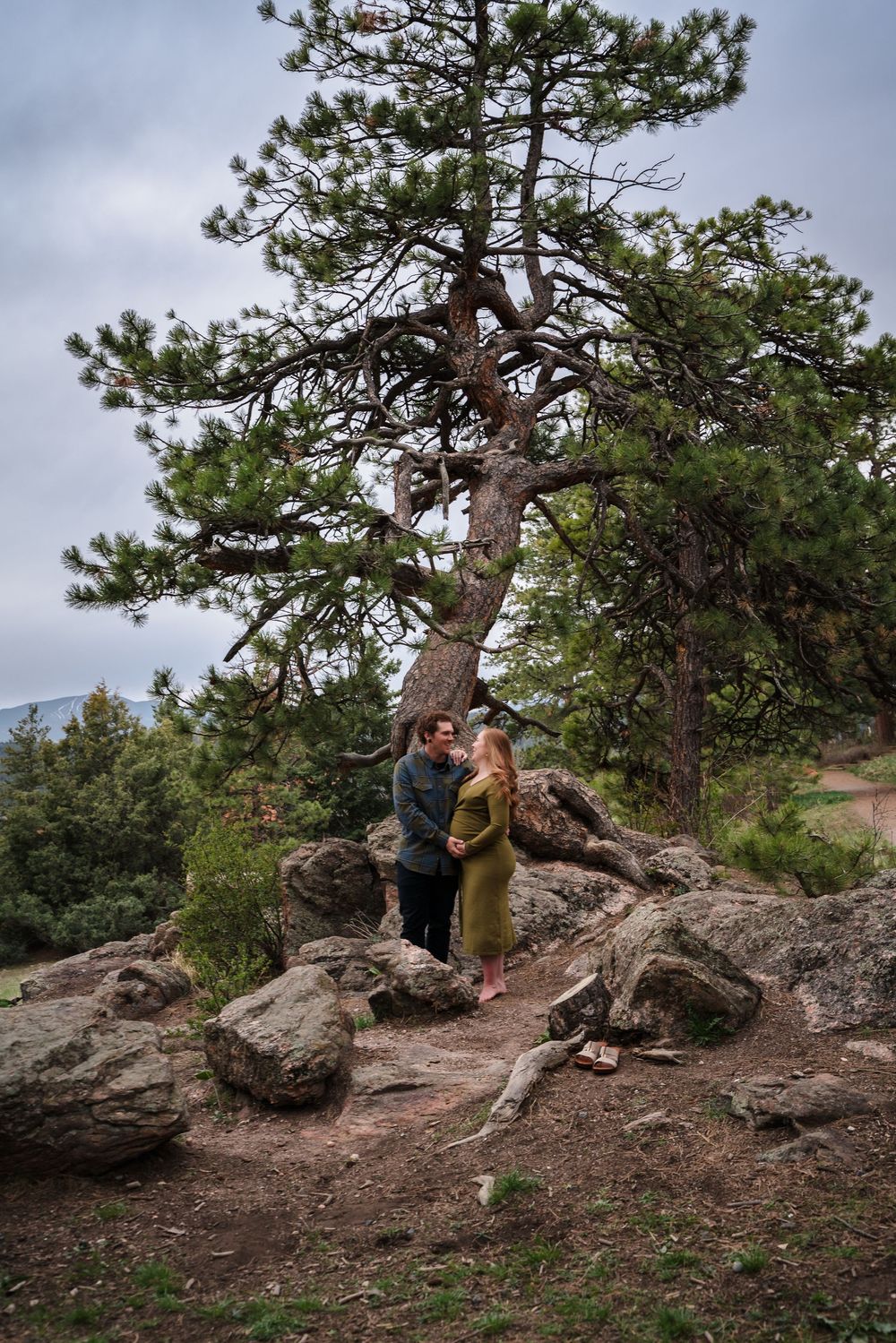 A couple poses together on rocky terrain with pine trees and mountains in the background during an outdoor photoshoot.