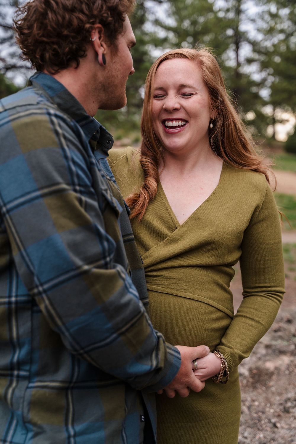 A couple shares intimate moments during a maternity photo shoot wearing olive green and plaid in an outdoor setting.