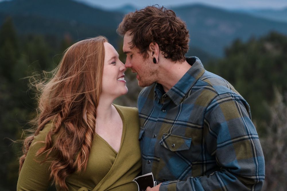 A romantic couple embraces outdoors with mountains and forests in the background during golden hour.