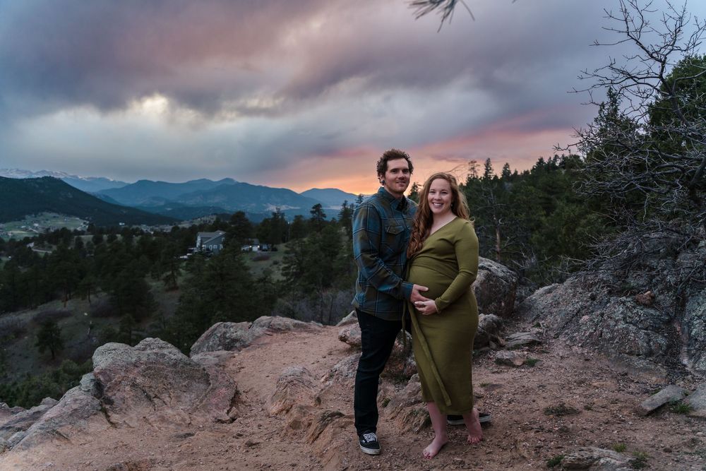 A couple embraces during sunset on a rocky mountain overlook with dramatic sky in the background.