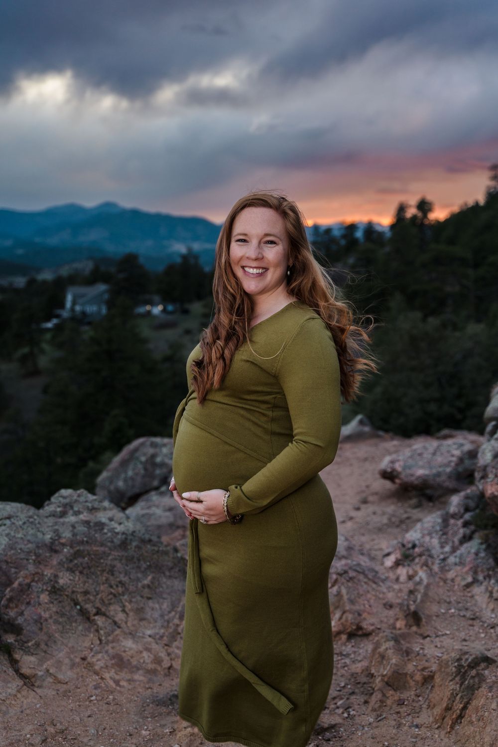 Woman in olive green dress cradles her pregnant belly against a dramatic mountain sunset backdrop.