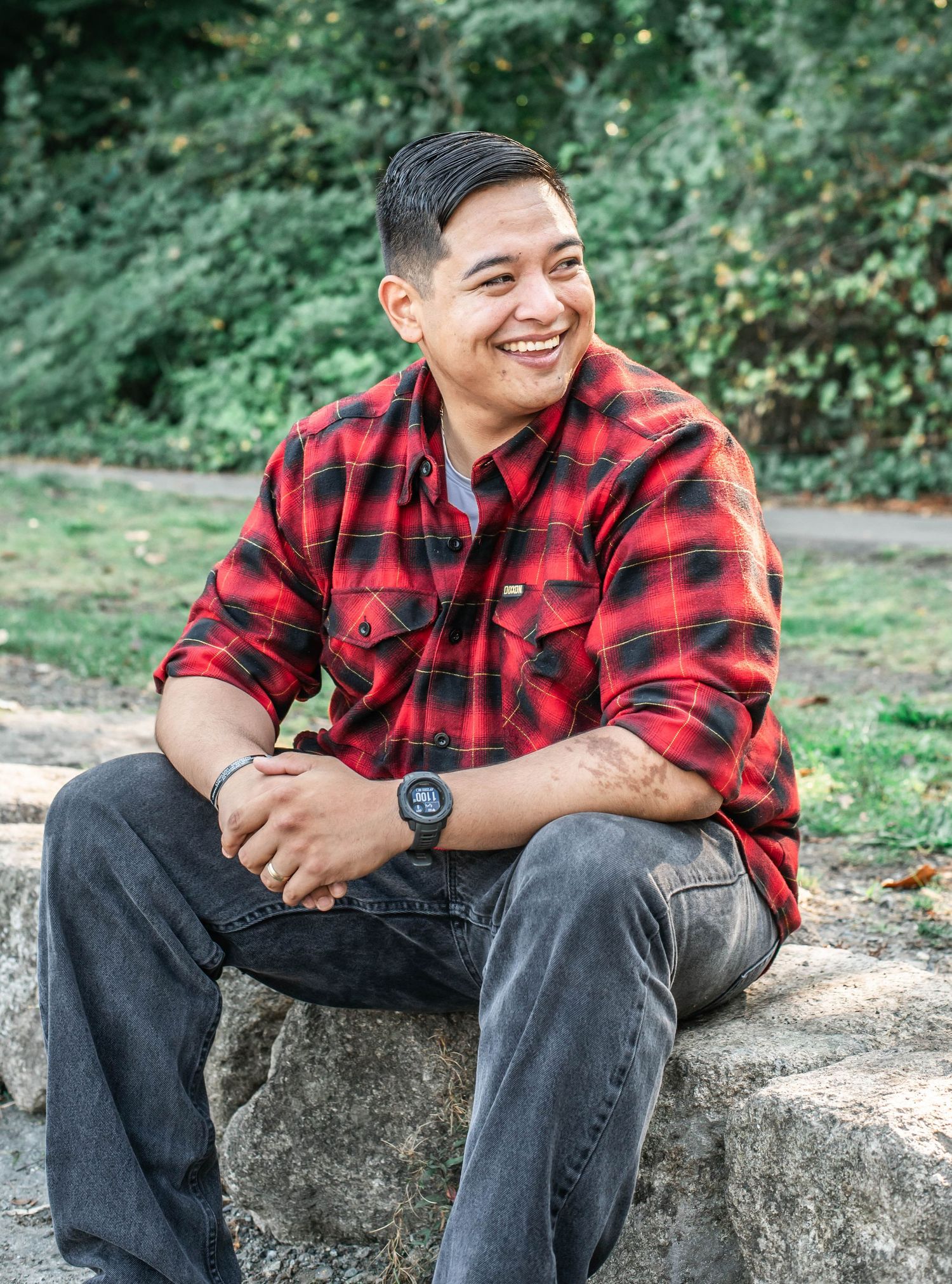 Person in red plaid shirt and jeans sitting casually on stone surface outdoors.