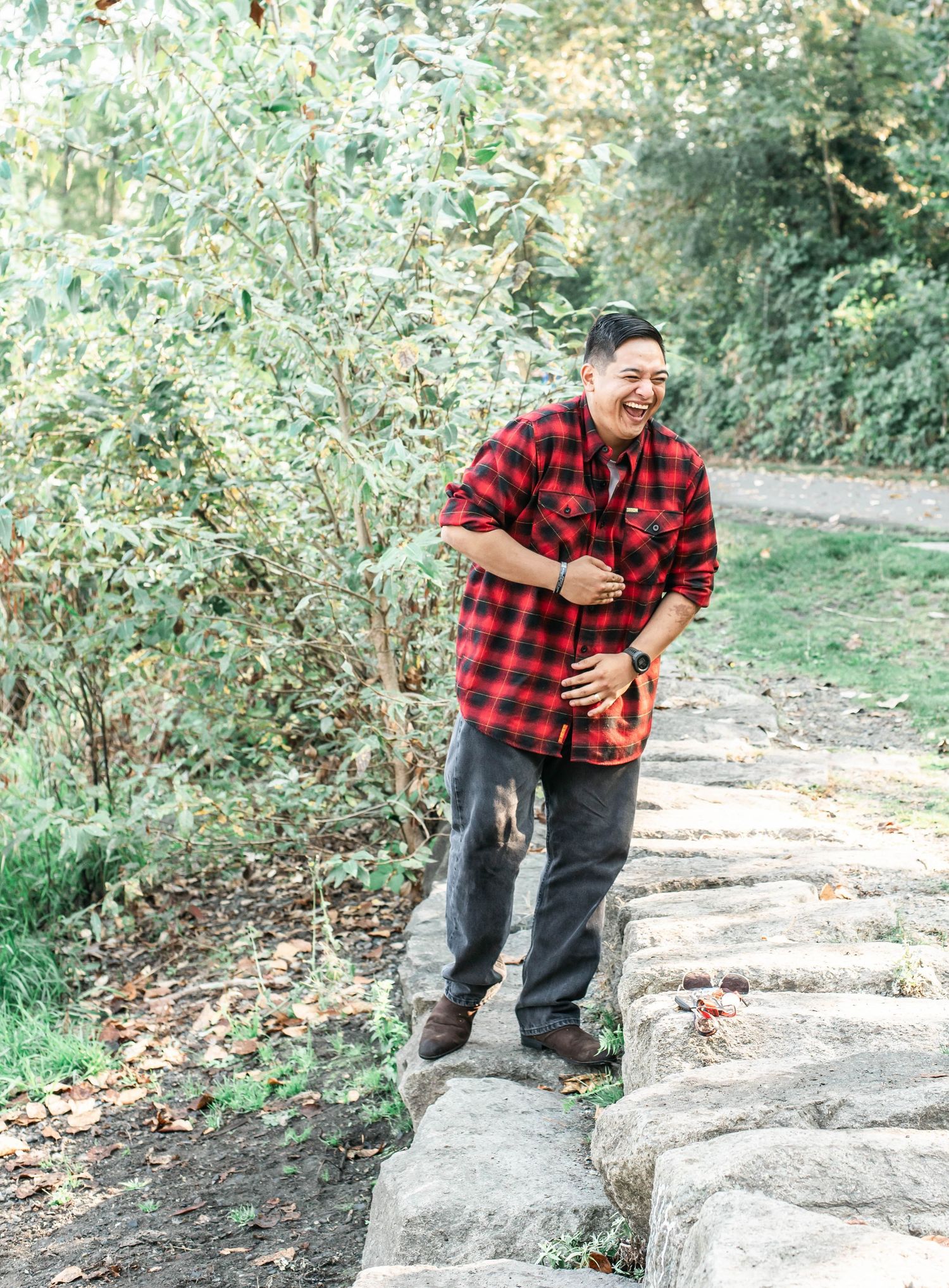 Person in buffalo plaid shirt poses playfully on natural stone steps in wooded setting.