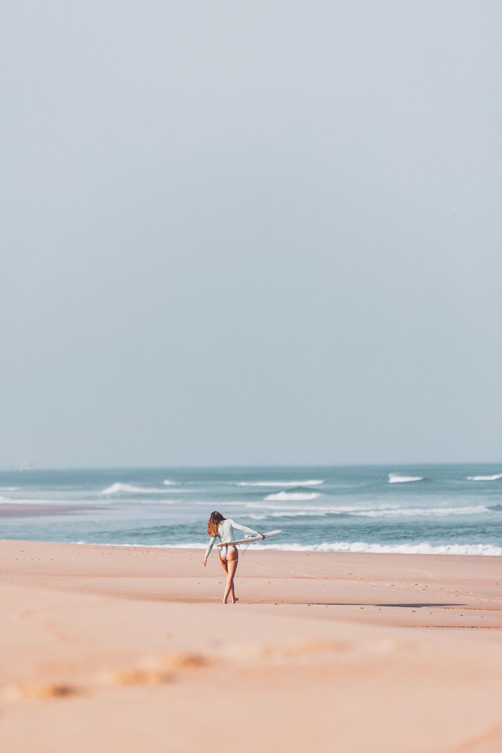 Une personne marche le long d'une plage sereine avec des vagues qui s'écrasent et du sable rose doux sous un ciel bleu
