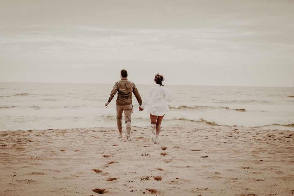 Deux personnages marchent main dans la main vers l'océan sur une plage brumeuse au coucher du soleil.