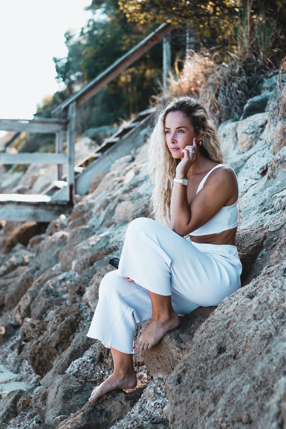 Une personne en robe blanche pose élégamment contre des falaises côtières rocheuses lors d'une séance photo au bord de mer au coucher du sol