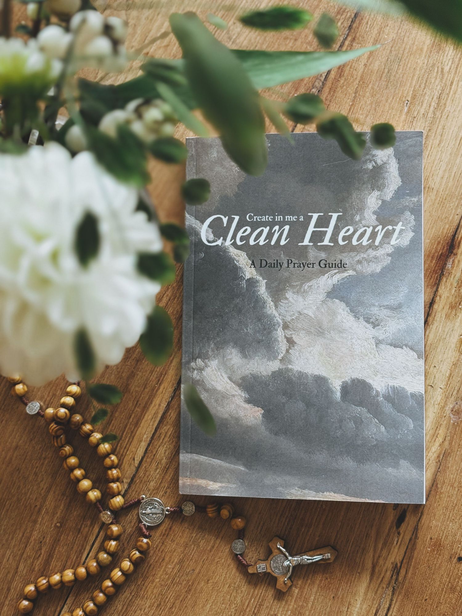 Prayer guide book titled 'Clean Heart' displayed with wooden rosary and white flowers on wooden surface.