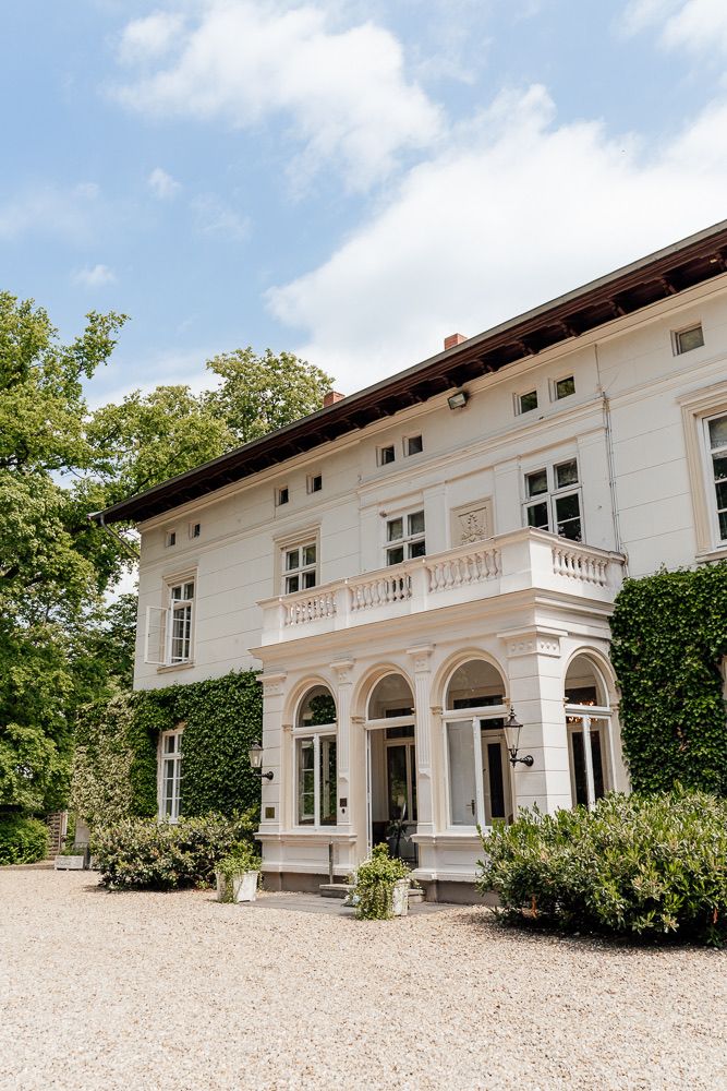 Elegant white Mediterranean-style villa with arched windows and balustrade details set against a blue sky and lush greenery.