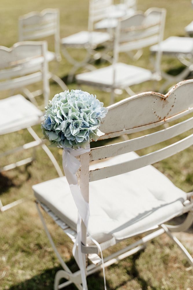 White folding chairs decorated with soft blue hydrangea flowers arranged for an outdoor wedding ceremony.